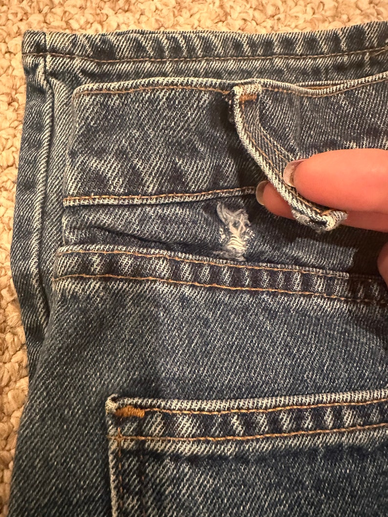 May include: Close-up of the back pocket of a pair of blue denim jeans. The pocket has a flap and is stitched with a contrasting thread.