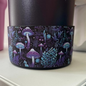 Puede incluir: Una taza reutilizable negra con un diseño colorido de hongos y follaje. La taza tiene una banda negra alrededor de la parte superior e inferior.