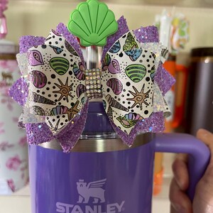 May include: A purple Stanley mug with a decorative bow. The bow features a seashell design with glitter accents and a green seashell charm. The bow is embellished with rhinestones.