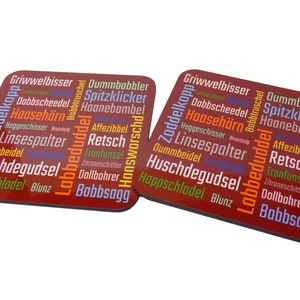 Könnte beinhalten: Zwei rote quadratische Untersetzer mit weißem Rand. Die Untersetzer sind mit einer Wortwolke aus deutschen Wörtern in verschiedenen Farben bedeckt, darunter Orange, Gelb, Blau und Lila. Die Wörter sind kreisförmig angeordnet.