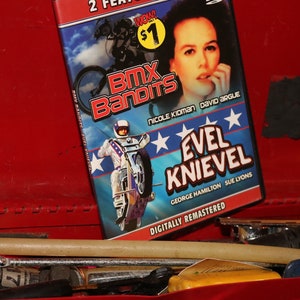 May include: A DVD case with a red, white, and blue design. The case features a photo of a woman and the text "BMX Bandits" and "Evel Knievel".