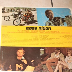 Puede incluir: Una portada de álbum de vinilo vintage para la película "Easy Rider" con Peter Fonda, Dennis Hopper y Jack Nicholson. La portada es amarilla con texto negro e imágenes de los actores y una motocicleta.