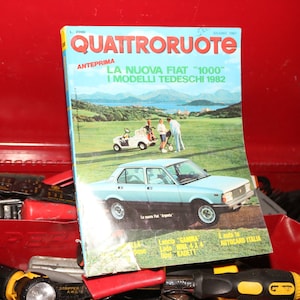 以下が含まれることがあります： 緑のゴルフコースに青いフィアット1000が載っている、ビンテージな雑誌の表紙。雑誌のタイトルは「クアトロルオテ」で、表紙のテキストは「ラ・ヌオバ・フィアット「1000」I モデルリ・テデスキ 1982」です。