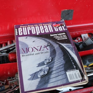 以下が含まれることがあります： 赤い工具箱の上に、「European Car」というタイトルの雑誌が置かれています。雑誌の表紙には、レース場の画像が掲載されており、「Monza Beautiful and Deadly」と「Racing Legends」の文字が書かれています。