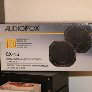 Puede incluir: Una caja blanca con texto negro y amarillo para un par de altavoces coaxiales Audiovox CX-15 de 15,2 cm. La caja muestra los altavoces con una potencia máxima de 120 vatios por par.