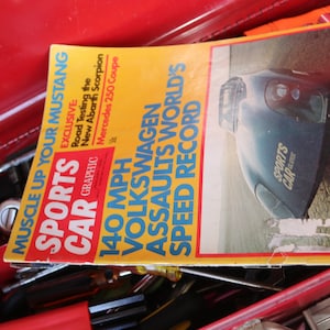 May include: A red toolbox with a magazine titled "Sports Car" on top. The magazine cover features a blue car and the text "140 MPH Volkswagen Assaults World's Speed Record".