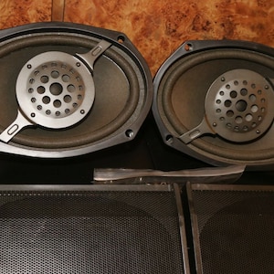 May include: Two oval-shaped car speakers with black plastic frames and a silver speaker grille. The speakers are sitting on a black surface.
