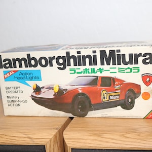 Puede incluir: Un coche de juguete rojo Lamborghini Miura en una caja con las palabras "lamborghini Miura" y "ランボルギーニミウラ" impresas en ella. La caja también dice "Real Action Head Lights", "Battery Operated", "Mystery Bump-N-Go Action", y "Chi Hung".