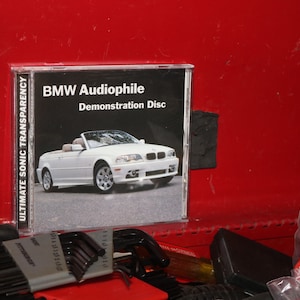 Puede incluir: Un coche descapotable BMW blanco aparece en la parte delantera de una caja de CD. La caja está etiquetada como "BMW Audiophile Demonstration Disc" y "Ultimate Sonic Transparency".