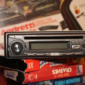 May include: A black car stereo with a silver control knob and a digital display. The stereo is sitting on top of a stack of VHS tapes. The tapes have titles like "Crashes" and "Magnum Force".