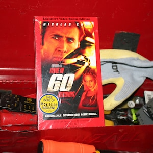 Könnte beinhalten: Eine rote VHS-Kassette des Films "Nur noch 60 Sekunden" mit Nicolas Cage und Angelina Jolie. Die Kassette befindet sich in einer roten Werkzeugkiste mit anderen Werkzeugen und einem Arbeitshandschuh.