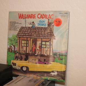 Könnte beinhalten: Ein Vinyl-Plattencover mit einer Cartoon-Illustration eines gelben Cadillac-Wagens, der vor einem kleinen Haus geparkt ist. Der Albumtitel lautet "Welfare Cadillac" von Guy Drake. Der Preisschild zeigt 4,98 $.