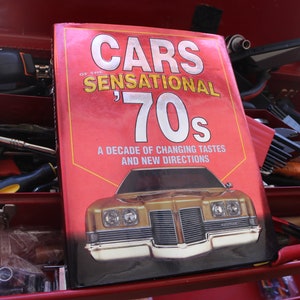 Peut inclure: Une boîte à outils rouge avec un livre intitulé "Cars of the Sensational '70s: A Decade of Changing Tastes and New Directions" sur le dessus. Le livre présente une voiture dorée sur la couverture.