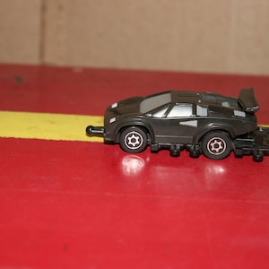 Puede incluir: Un coche de juguete negro con un alerón y ruedas. El coche está sobre una superficie roja y amarilla.