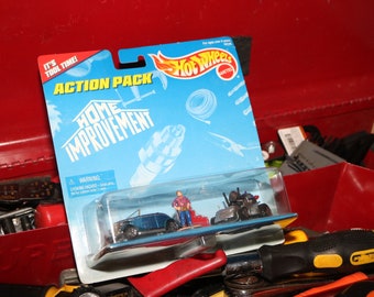 Könnte beinhalten: Ein blaues und rotes Hot Wheels Action Pack mit dem Thema Heimwerken. Das Pack enthält ein blaues Auto, einen roten LKW und eine Figur eines Mannes, der ein blaues Hemd und Jeans trägt. Die Verpackung trägt die Aufschrift "It's Tool Time!" und "Home Improvement".