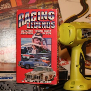 Peut inclure: Une cassette VHS rouge avec le titre "Racing Legends" présentant les noms de Joe Weatherly, Fireball Roberts, Curtis Turner et Tim Flock. La jaquette de la cassette montre une voiture de course vintage avec les mots "Fabulous Hudson Hornet" sur le côté.