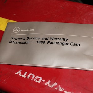 May include: A grey pamphlet with the text "Mercedes-Benz Owner's Service and Warranty Information - 1999 Passenger Cars" on a red surface.