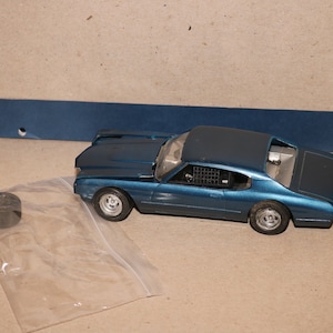 Puede incluir: Un coche modelo azul con interior negro y jaula antivuelco. El coche está sobre una superficie marrón con una bolsa de plástico y un neumático negro.