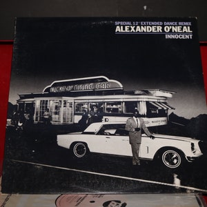 May include: A black and white vinyl record cover featuring a black and white photo of a man standing next to a white car in front of a diner. The diner has a sign that reads "Mickey's Dining Car". The record cover is for the song "Innocent" by Alexander O'Neal.
