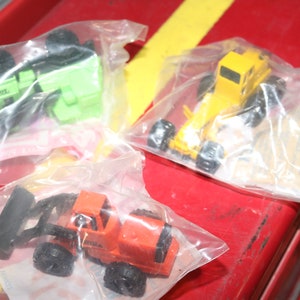 May include: Four toy construction vehicles in individual plastic bags. The vehicles are a green bulldozer, a yellow dump truck, a red front loader, and an orange backhoe.