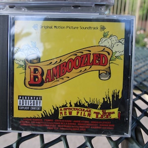 May include: A yellow CD case with a black parental advisory sticker. The case features a black and white image of a crowd and a banner that reads "A Spectacular New Film by Spike Lee". The title of the soundtrack is "Bamboozled" in red and yellow lettering.