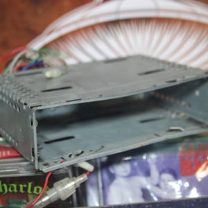 Puede incluir: Un kit de montaje para radio de coche de metal gris, de forma rectangular con un recorte en el centro. El kit tiene una serie de pequeños agujeros a lo largo de los bordes.