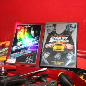 May include: Two DVD cases for the movies "Fast & Furious" and "2 Fast 2 Furious". The cases are red and black with images of cars and actors on the front.