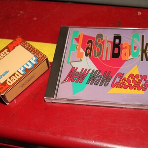 May include: A CD case with the text "FlashBack! New Wave Classics" in a colorful design. Beside it is a small gift box with the words "father pop" and "dad pup" printed on it. The items are on a red surface.