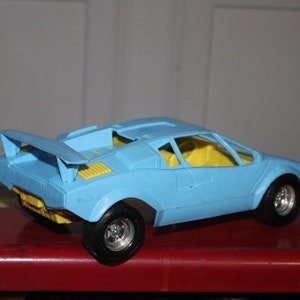Puede incluir: Un coche de juguete azul claro con interior amarillo y alerón trasero. El coche de juguete tiene ruedas negras con llantas plateadas. El coche está sobre una superficie roja con rayas amarillas.