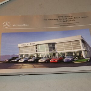 May include: A 1998 directory for Mercedes-Benz passenger car and light truck dealers in the United States and Canada. The cover features a photo of a modern glass and steel dealership building with a row of Mercedes-Benz cars parked in front.