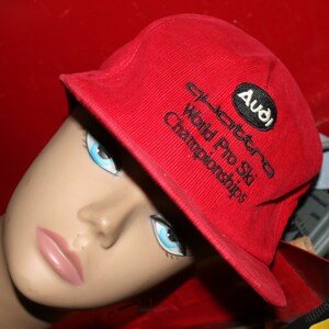 May include: A red baseball cap with the text "Audi quattro World Pro Ski Championships" embroidered on the front. The cap is worn by a mannequin with blue eyes and a pink lip.