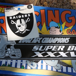 May include: Two Raiders stickers, one with the team logo and the words "RAIDERS" and the other with a helmet and the words "2002 AFC CHAMPIONS SUPER BOWL XXXVII".