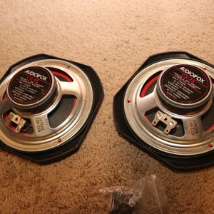 May include: Two AudioVox CX-15 car speakers with black surrounds, silver cones, and red accents. Each speaker has a central black label with white text. The speakers are shown on a brown surface.