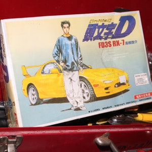 Könnte beinhalten: Ein gelbes und schwarzes Modellauto mit einer blauen und weißen Schachtel. Die Schachtel trägt den Text "Initial D FD3S RX-7". Das Auto ist ein Mazda RX-7.