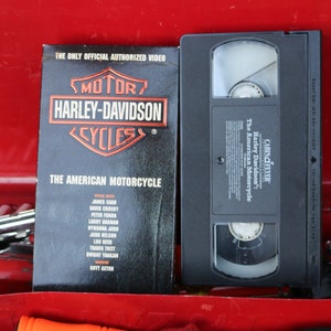 May include: A VHS video cassette tape with a black label and the text "CABIN FEVER The American Motorcycle Harley Davidson" on it. The tape is in a red toolbox with a black book that says "THE ONLY OFFICIAL AUTHORIZED VIDEO MOTOR HARLEY-DAVIDSON CYCLES THE AMERICAN MOTORCYCLE" on it.