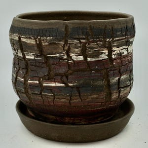 May include: A handcrafted ceramic planter with a textured, bark-like appearance. The pot features a dark brown rim and base, with layers of black, white, and red-brown hues. The planter sits on a matching saucer, suitable for indoor or outdoor use.