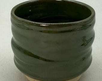 May include: A handmade, dark green ceramic cup with a textured, ridged design. The cup has a smooth, glossy finish and a light beige base. The interior is a darker shade of green. This unique piece is perfect for enjoying your favorite beverage.