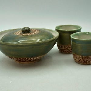 Może przedstawiać: Ręcznie wykonany ceramiczny zestaw do herbaty w odcieniach zieleni i brązu. Zestaw zawiera misę z pokrywką i teksturowanym brzegiem oraz dwa pasujące kubki. Misa ma dzióbek i ozdobny guzik na pokrywce.