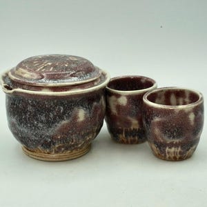 Może przedstawiać: Ręcznie wykonany zestaw ceramiczny składający się z dzbanka z pokrywką i dwóch małych filiżanek. Ceramika ma nakrapianą, ciemnobordową glazurę z kremowymi akcentami. Dzbanek ma zaokrąglony kształt z małym dziobkiem.