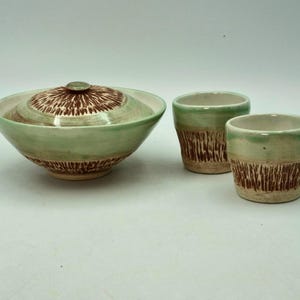 Könnte beinhalten: Eine handgefertigte Keramik-Servierschüssel mit Deckel und zwei passenden Tassen. Die Schüssel und die Tassen sind hellgrün mit braunen strukturierten Designs. Die Schüssel hat einen Deckel mit einem braunen strukturierten Muster.
