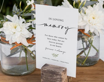May include: A white card with black text reads "In Loving Memory" and "For those who cannot be here today, but are forever present in our hearts forever + always". The card is displayed on a wooden stand with three white flowers in glass jars.