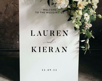 Peut inclure: Une pancarte de bienvenue blanche avec du texte noir qui dit "Bienvenue au mariage de Lauren et Kieran 22.09.22". La pancarte est décorée de fleurs blanches et de verdure.