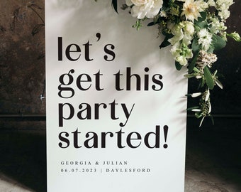 以下が含まれることがあります： 白い看板に黒い文字で「let's get this party started!」と書かれており、その下にGeorgia & Julianの名前と日付06.07.2023 | Daylesfordが書かれている。看板は白い花と緑の葉で飾られている。