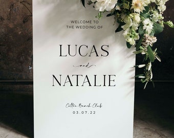 以下が含まれることがあります： 白い結婚式の看板に黒い文字で「LucasとNatalieの結婚式へようこそ、Cotton Beach Club、03.07.22」と書かれています。看板は白い花と緑の葉で飾られています。
