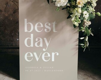 以下が含まれることがあります： 薄い茶色の看板に白い文字で「best day ever」と書かれている。テキストの下には「Georgia & Julian 06.07.2023 | Daylesford」という情報が書かれている。看板は白い花と緑の葉のブーケで飾られている。