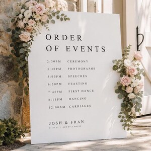 May include: A white rectangular wedding order of events sign with black serif text listing times and activities from ceremony to carriages. The sign is decorated with two floral arrangements of pale pink roses and green eucalyptus leaves.