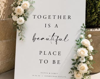 May include: A rectangular white sign featuring the black text TOGETHER IS A BEAUTIFUL PLACE TO BE, OLIVES & JAMES, and 10.06.26 | GOSBOURNE ESTATE. The sign is decorated with two clusters of cream-colored roses and green foliage on the top left and right.