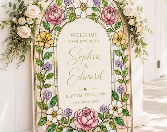 Op de afbeelding: Een boogvormig welkomstbord voor een bruiloft met een glas-in-loodrand van roze, gele en witte bloemen en groene bladeren. De tekst in het midden luidt: WELCOME TO OUR WEDDING, Sophia & Edward, SEPTEMBER 12, 2026, THE ORANGERY. Bloemstukken flankeren de bovenkant.