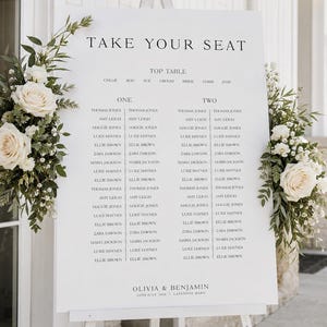 May include: A white wedding seating chart sign on an easel with the text TAKE YOUR SEAT, TOP TABLE, ONE, TWO, and OLIVIA & BENJAMIN. The sign is decorated with two floral arrangements featuring cream-colored roses and green foliage on the sides.