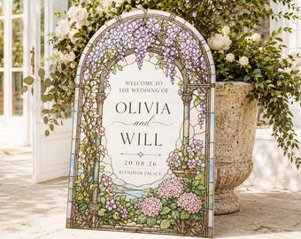 Op de afbeelding: Een boogvormig welkomstbord voor een bruiloft met een glas-in-loodontwerp van paarse blauweregen, roze hortensia's en groen blad. De tekst luidt: WELCOME TO THE WEDDING OF OLIVIA and WILL 20.08.26 BLENHEIM PALACE, tegen een landschapsachtergrond.